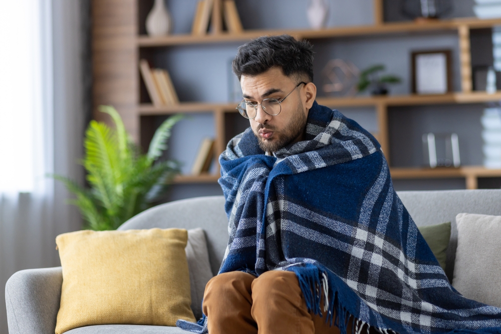 Man,wrapped,in,checkered,blanket,sitting,on,sofa,,looks,cold